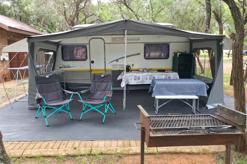 A Jurgens Penta caravan on a paved stand beside a brick house, awning out, with two camp chairs, a table and a braai