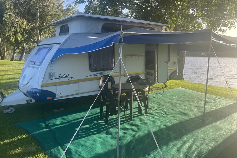 A pop-top caravan set up beside the water with its awning out and chairs under it
