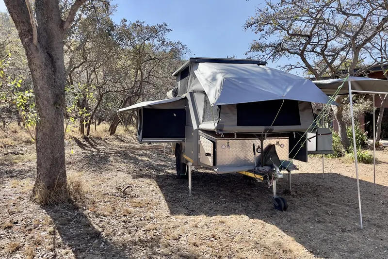 A Compass Seekoei off-road caravan reversed onto a bush stand between the trees, with its pop-top up and an awning out