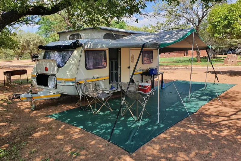 A Jurgens Penta pop-top caravan under trees with a groundsheet, camp chairs and a cooler box